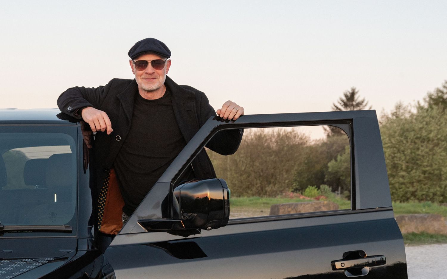 Man in black outfit and cap leaning on open door of dark pickup truck outdoors at dusk.