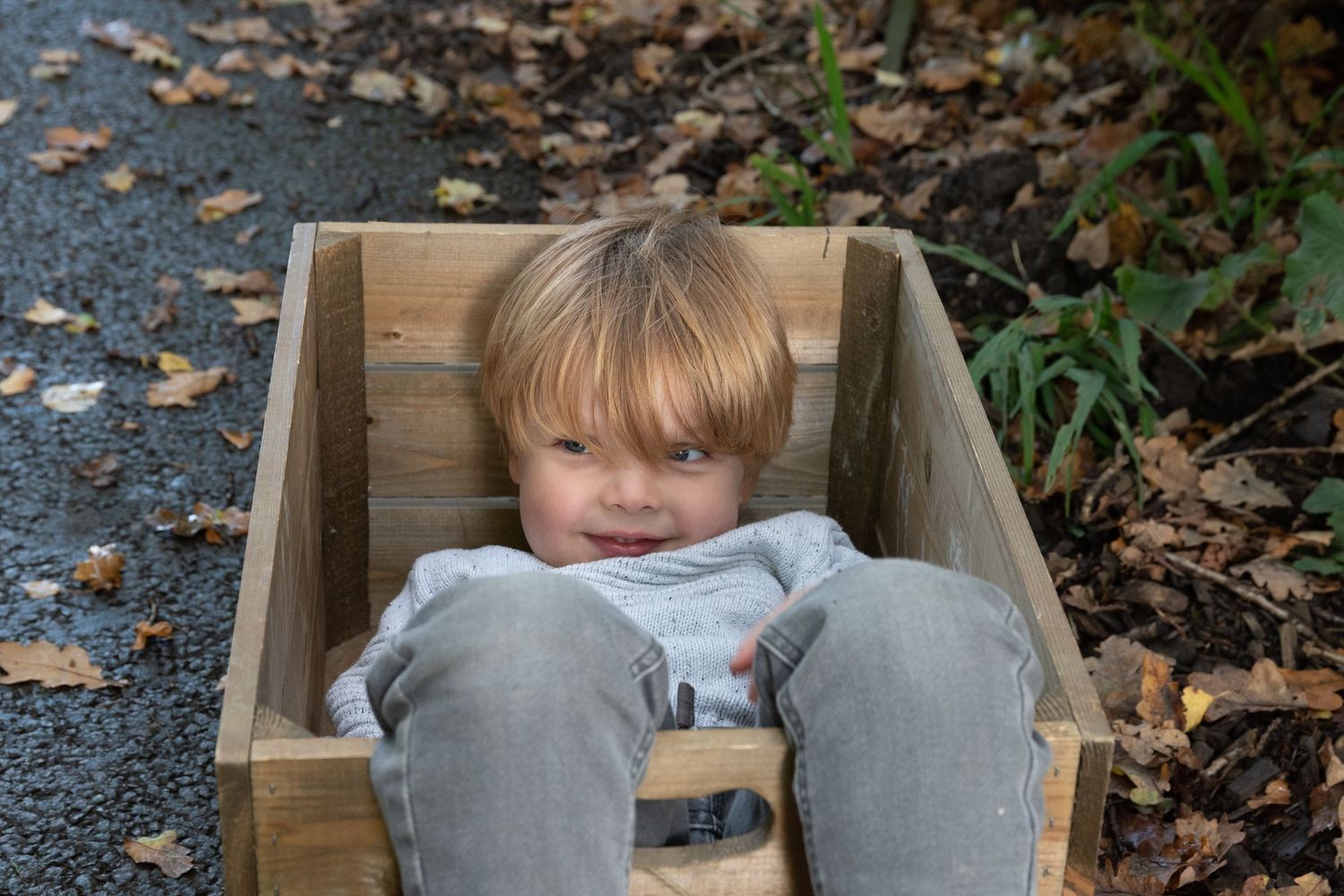 A small child sits inside a wooden wagon on a path covered with autumn leaves.