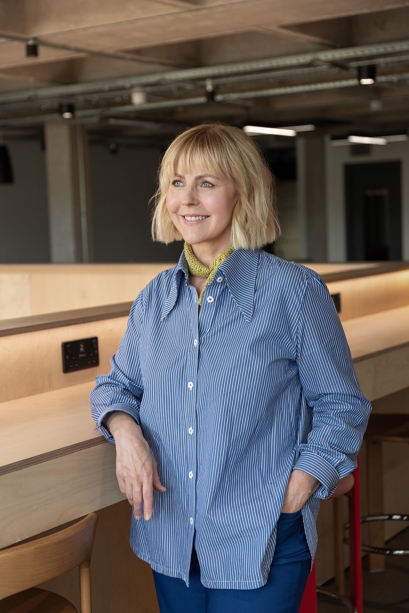 Smiling blonde woman in blue striped shirt leaning against wooden counter in modern office space.