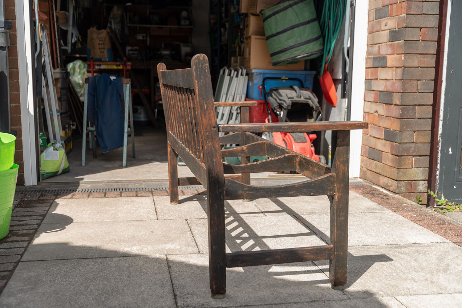 Weathered teak garden bench on a sunny patio outside a garage filled with outdoor equipment.
