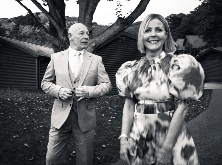 A man in a suit and a woman in a floral dress smile outdoors near trees and buildings in a black and white photo.