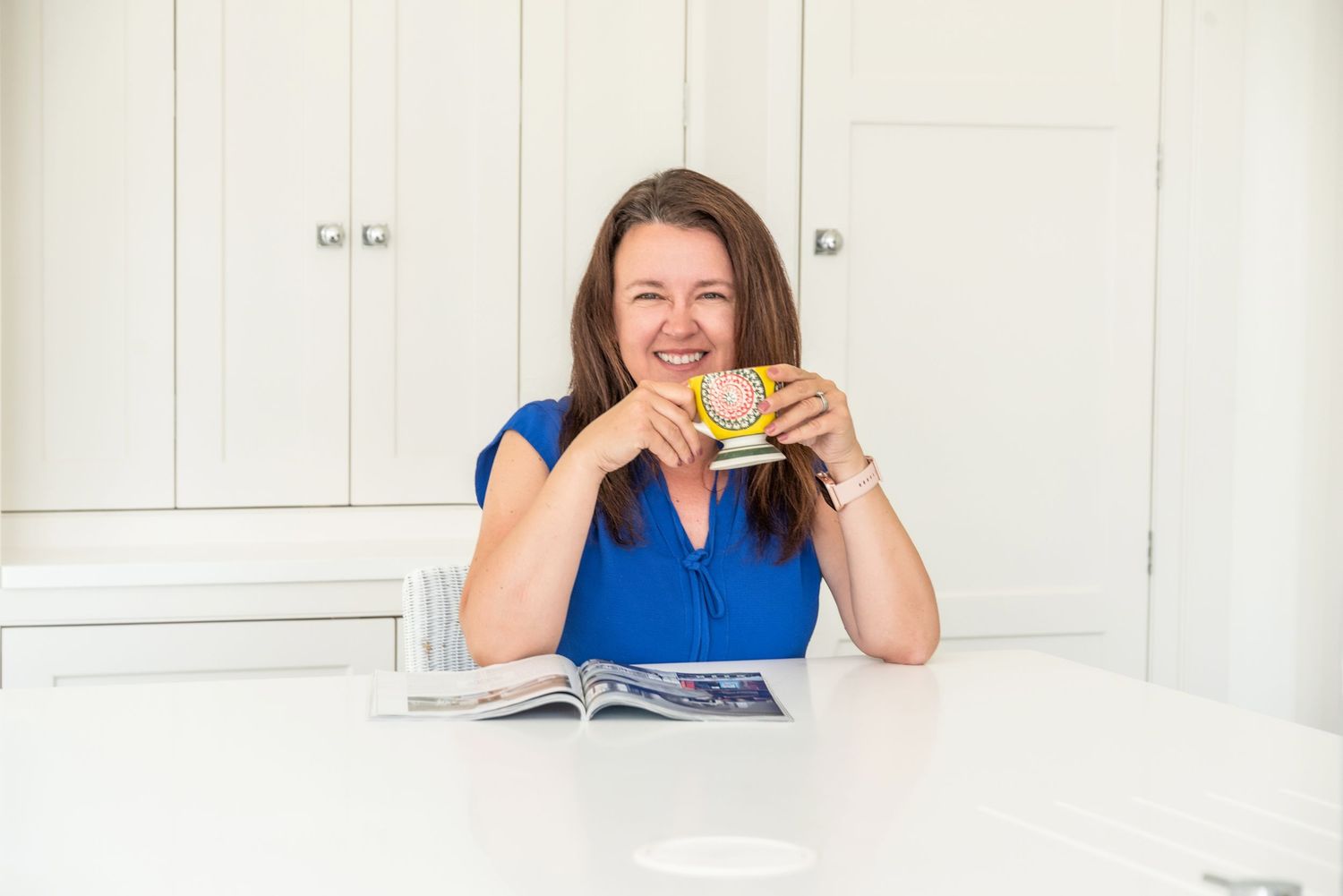 Woman in blue top laughing while holding yellow mug at white kitchen table with open magazine.