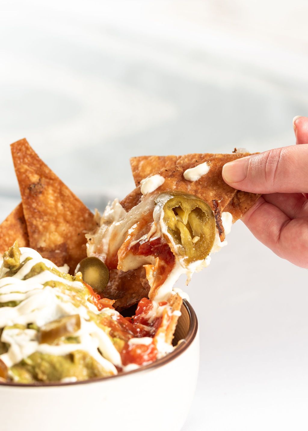 A hand holding a loaded nacho chip topped with jalapenos, sour cream, and melted cheese over a bowl of nachos.