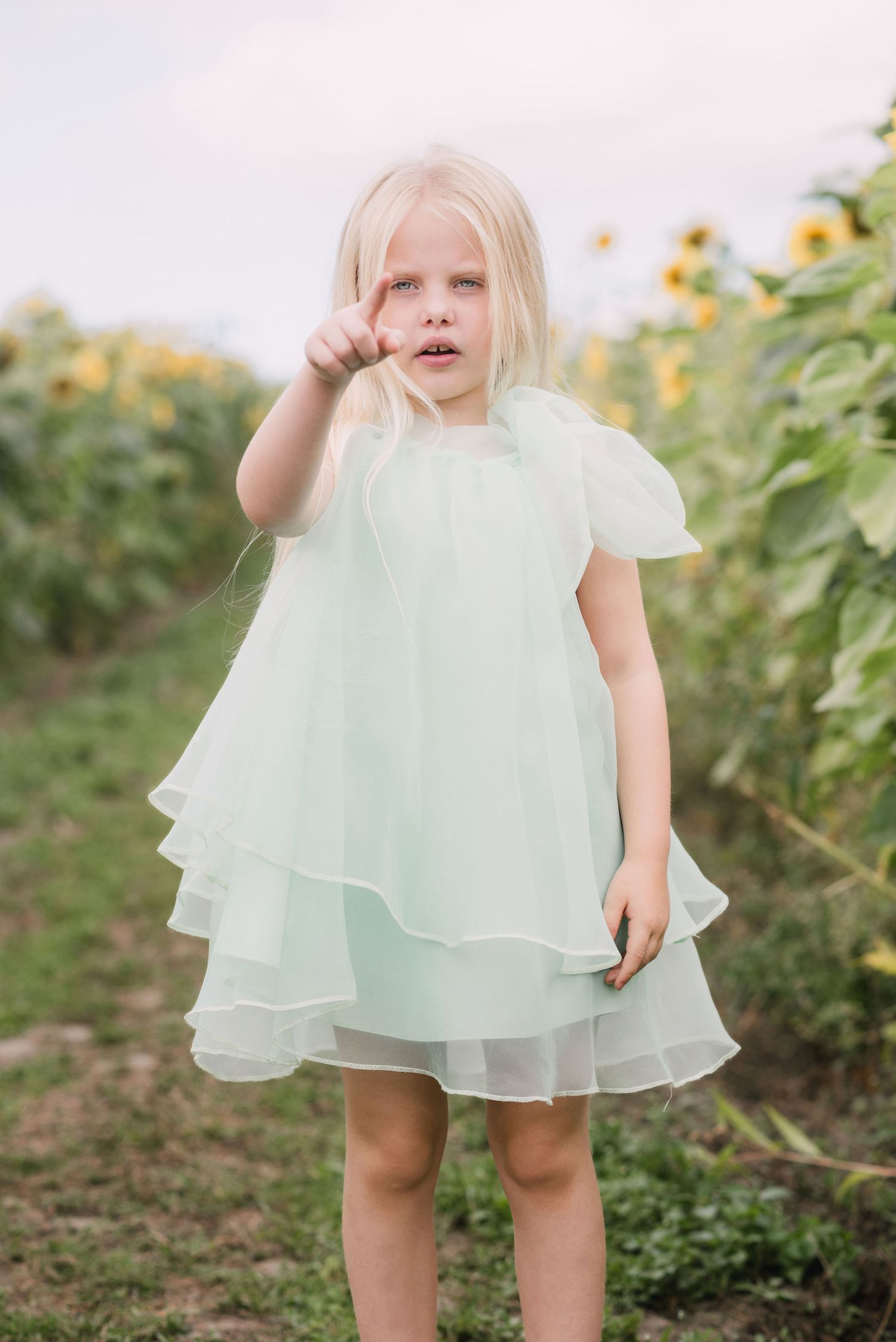 A child wearing a mint green ruffled dress twirls in a garden with sunflowers in the background during golden hour.
