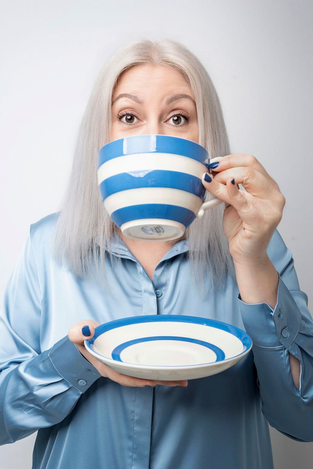 Woman with white hair drinking from a large blue and white striped cup and saucer, wearing a blue shirt.