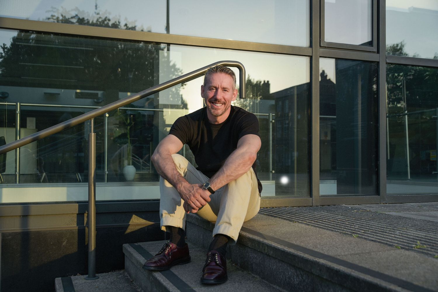 Smiling man sitting on steps outside a modern glass building, wearing a black t-shirt and khaki pants.
