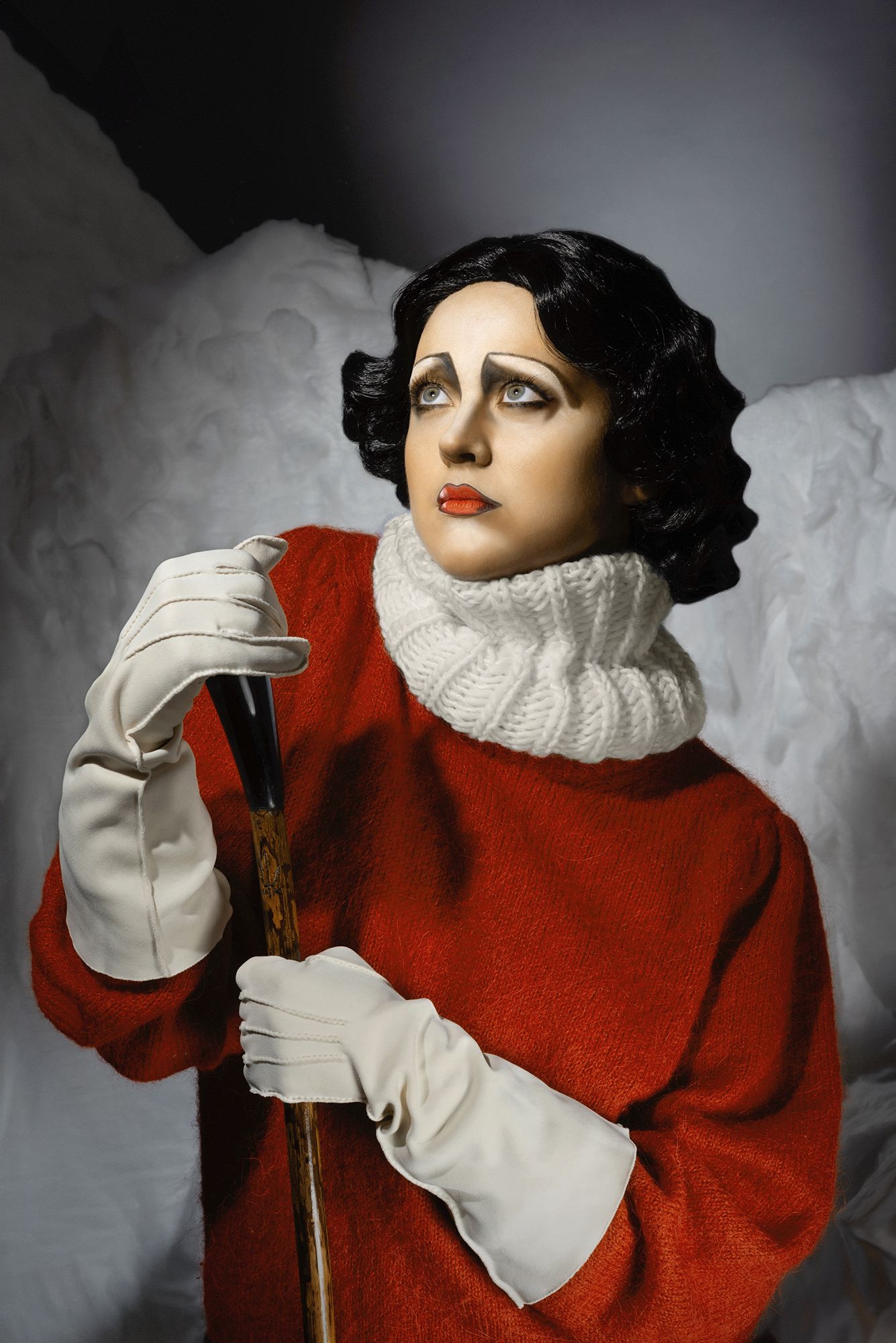 Dramatic portrait of figure in red coat with white turtleneck and gloves against dark background showcasing vintage glamour.
