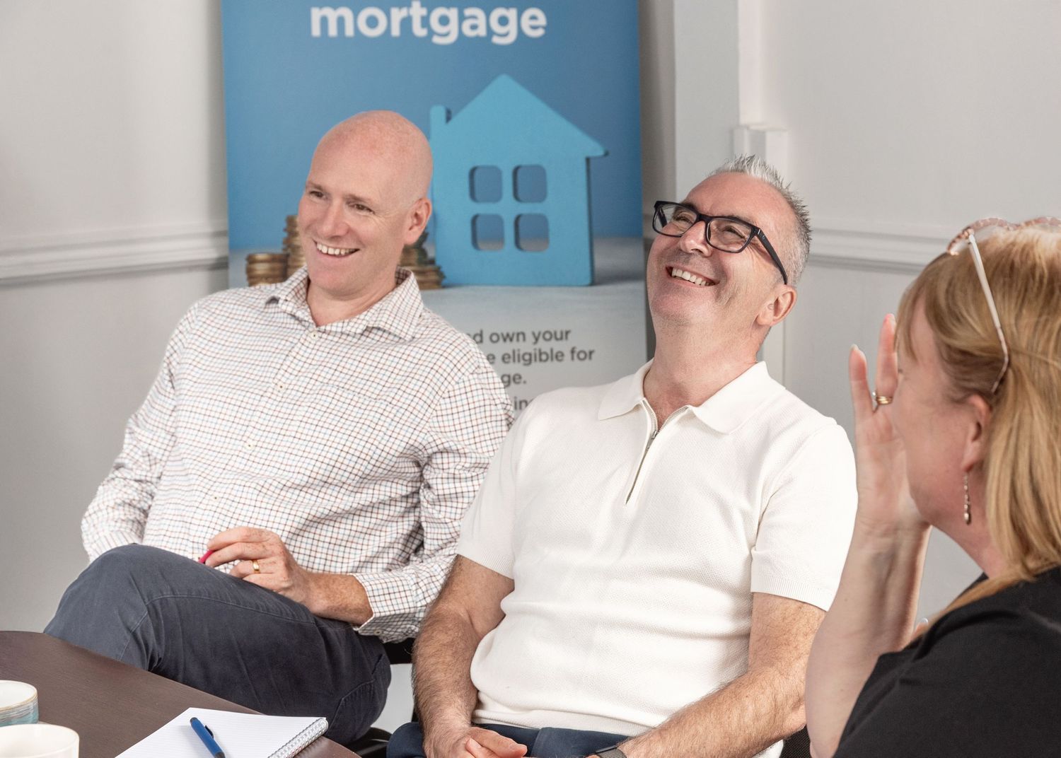 People share a lighthearted moment during a business meeting with a blue house mortgage logo displayed behind them.