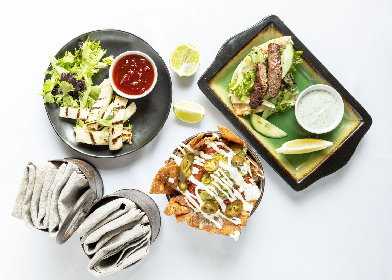 Overhead view of Mexican food spread with nachos, grilled meat platter, salsa dip, tortillas, and fresh garnishes on white background.