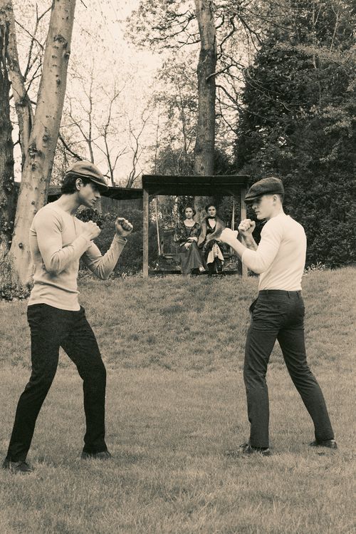 Two men in vintage caps spar in a sepia-toned outdoor scene with a framed portrait of women between them.