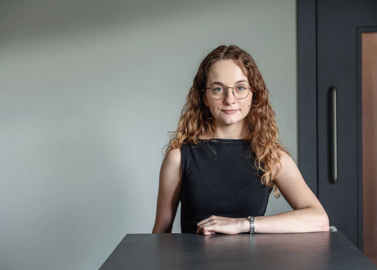 Young woman with curly red hair and glasses sitting at a table in a modern office setting.