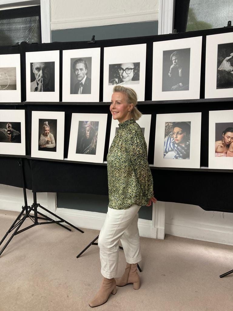 Woman in green floral blouse and white pants standing in front of black and white portrait photography exhibition.