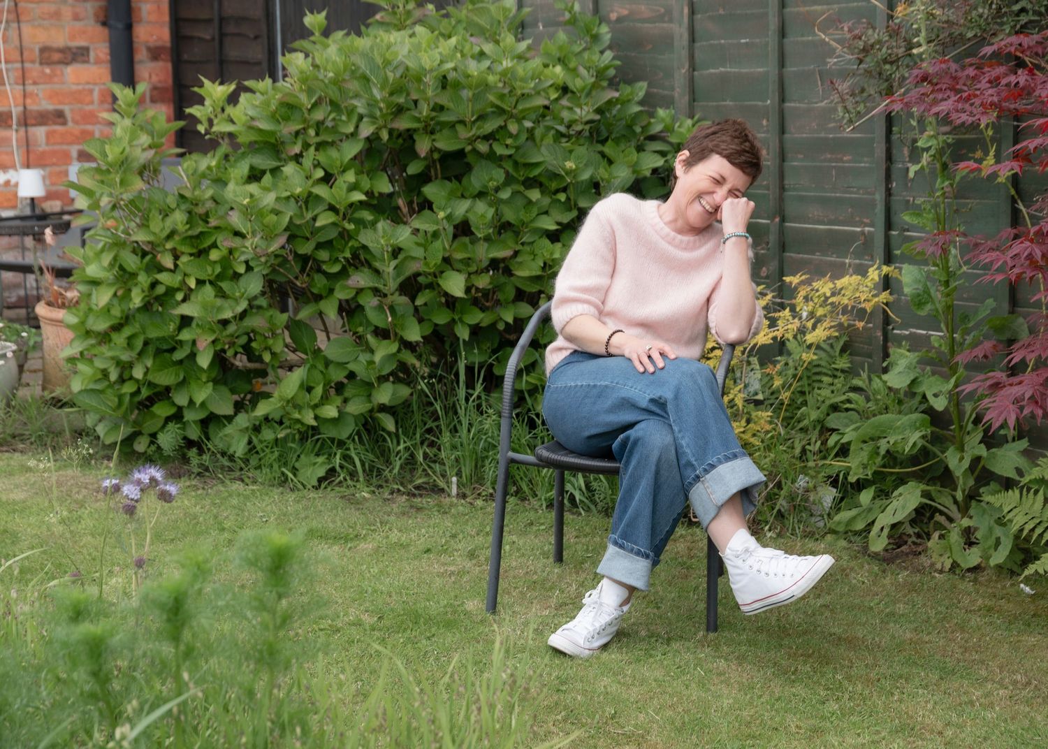 Woman in pink sweater and jeans sits on garden chair outdoors, surrounded by lush green shrubs and plants.