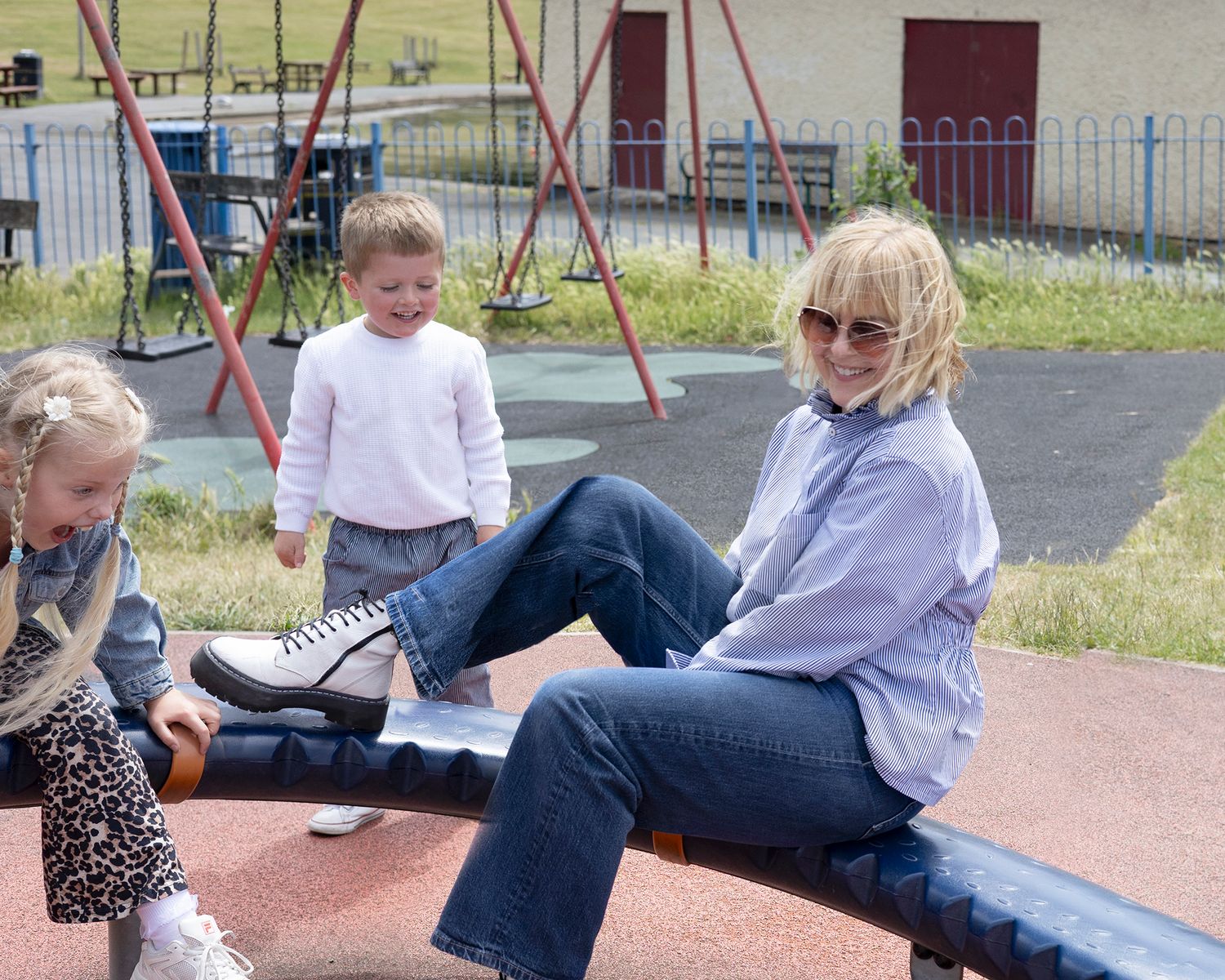 Woman in jeans sits on seesaw with two young children at outdoor playground on sunny day.