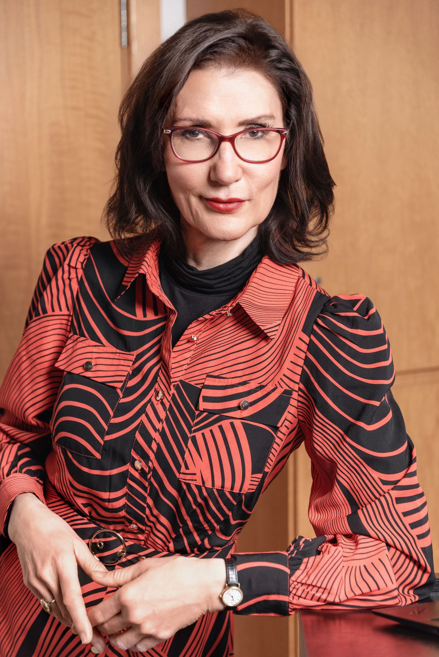 Professional portrait showing someone wearing glasses and a stylish red and black patterned blouse with long sleeves.