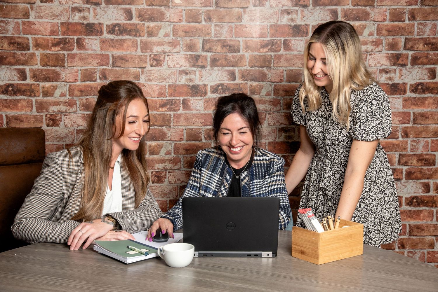Three colleagues laugh while looking at laptop screen during casual meeting at rustic brick wall office space.
