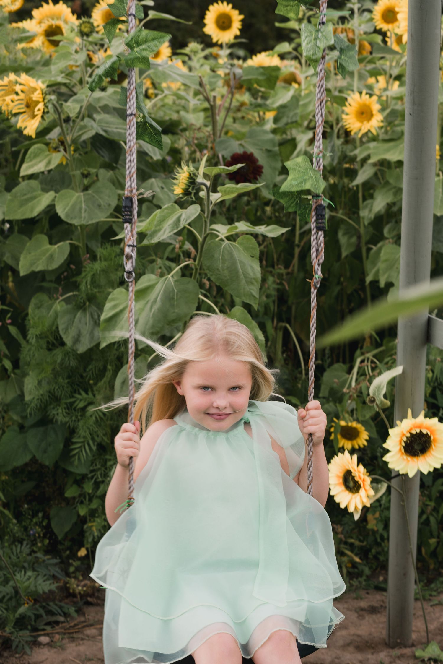 A child on a swing set in front of a vibrant garden of tall blooming sunflowers.