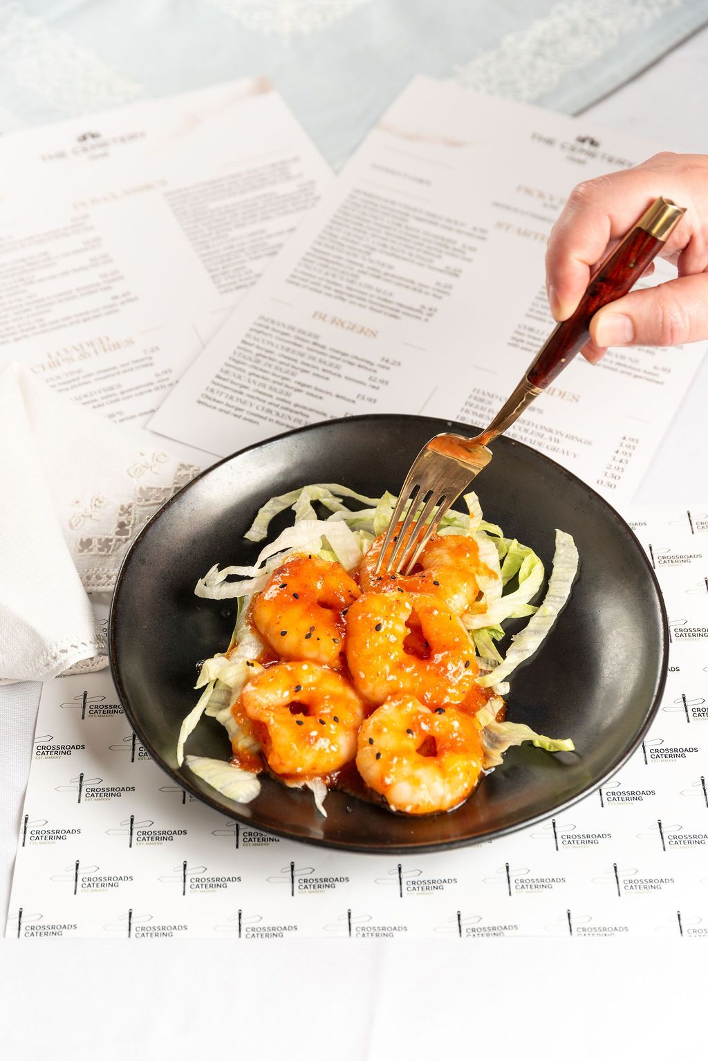 Glazed shrimp on shredded cabbage in a dark bowl, with a hand holding a fork, menus in the background.