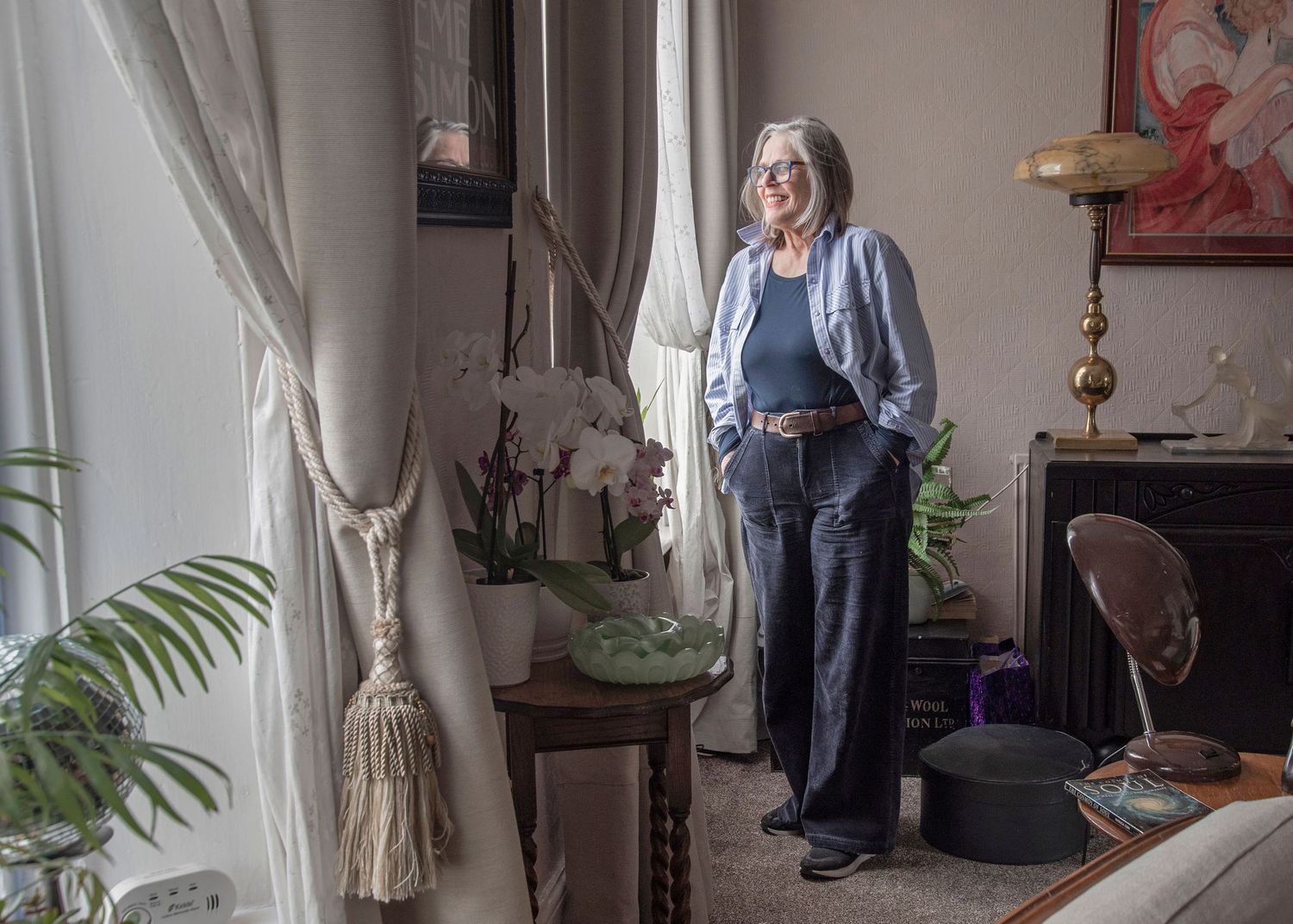 Elegant older woman with gray hair posing by window in stylish home with orchids and vintage decor.