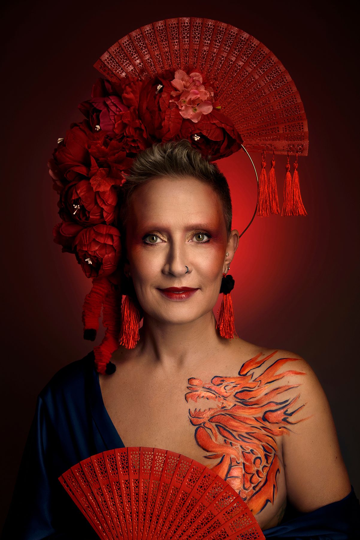 Artistic portrait with red fan headdress, blue dress and phoenix body art against dark red background.