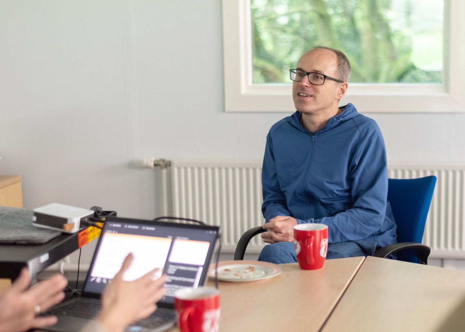 Someone in glasses and blue sweater participates in meeting discussion from across office desk with colleague.