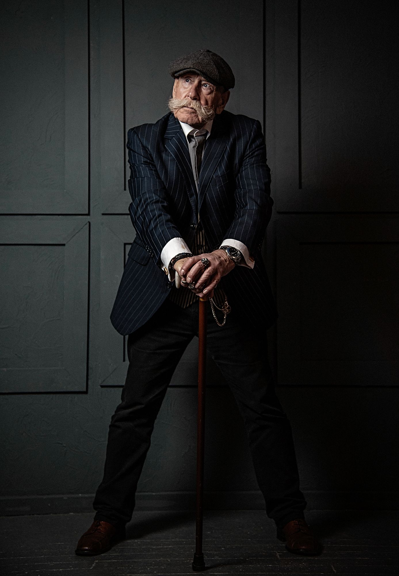 Dramatic portrait of person in pinstripe suit and cap holding cane in dark moody setting.