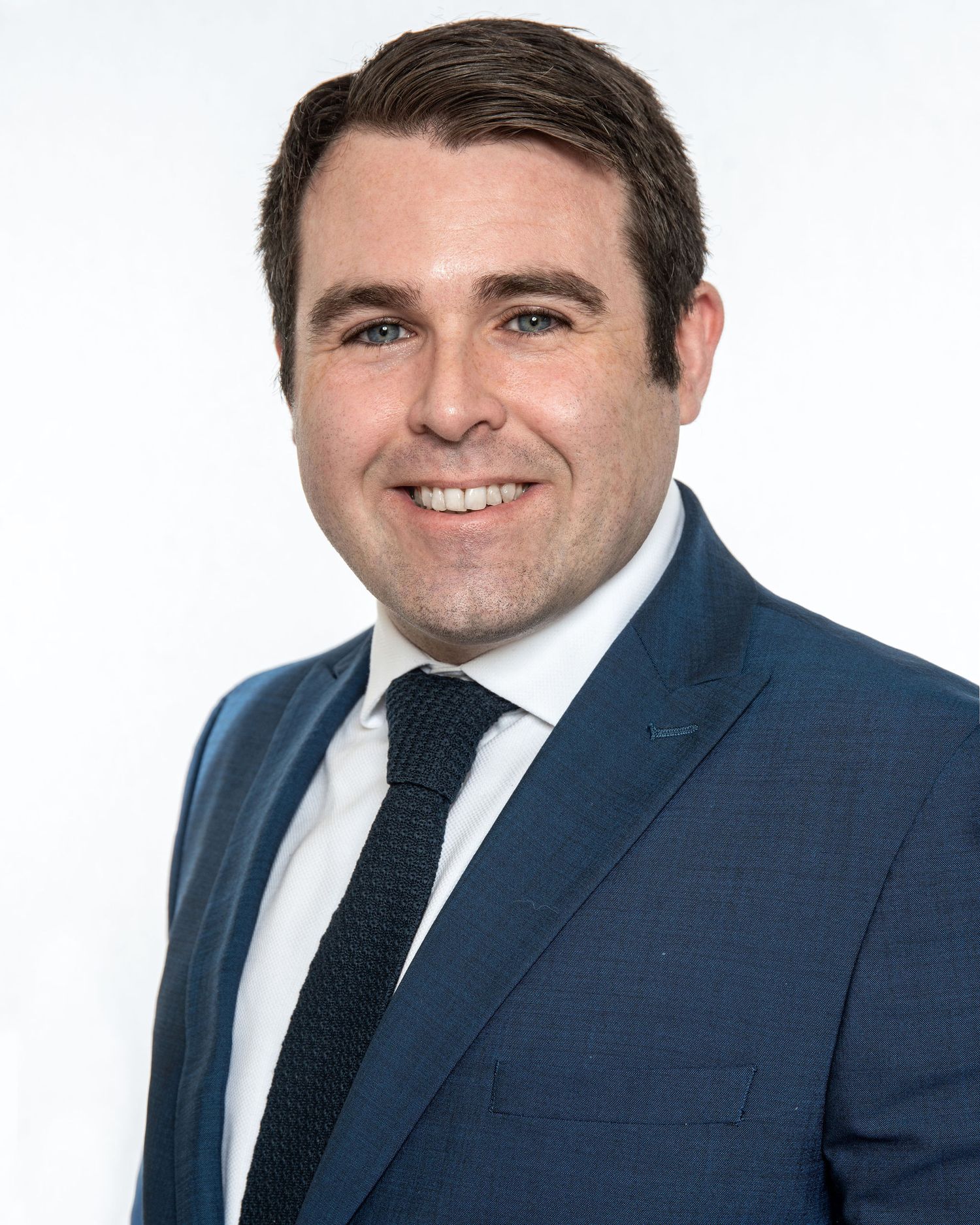 Professional headshot of person in navy suit and tie against white background.