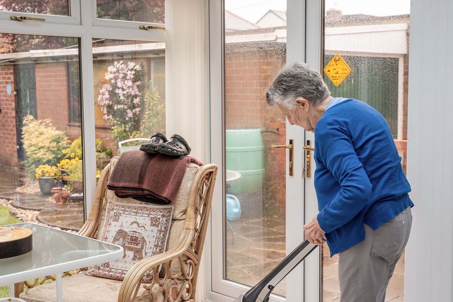 Elderly woman vacuuming a bright conservatory with garden views through large glass windows and doors.