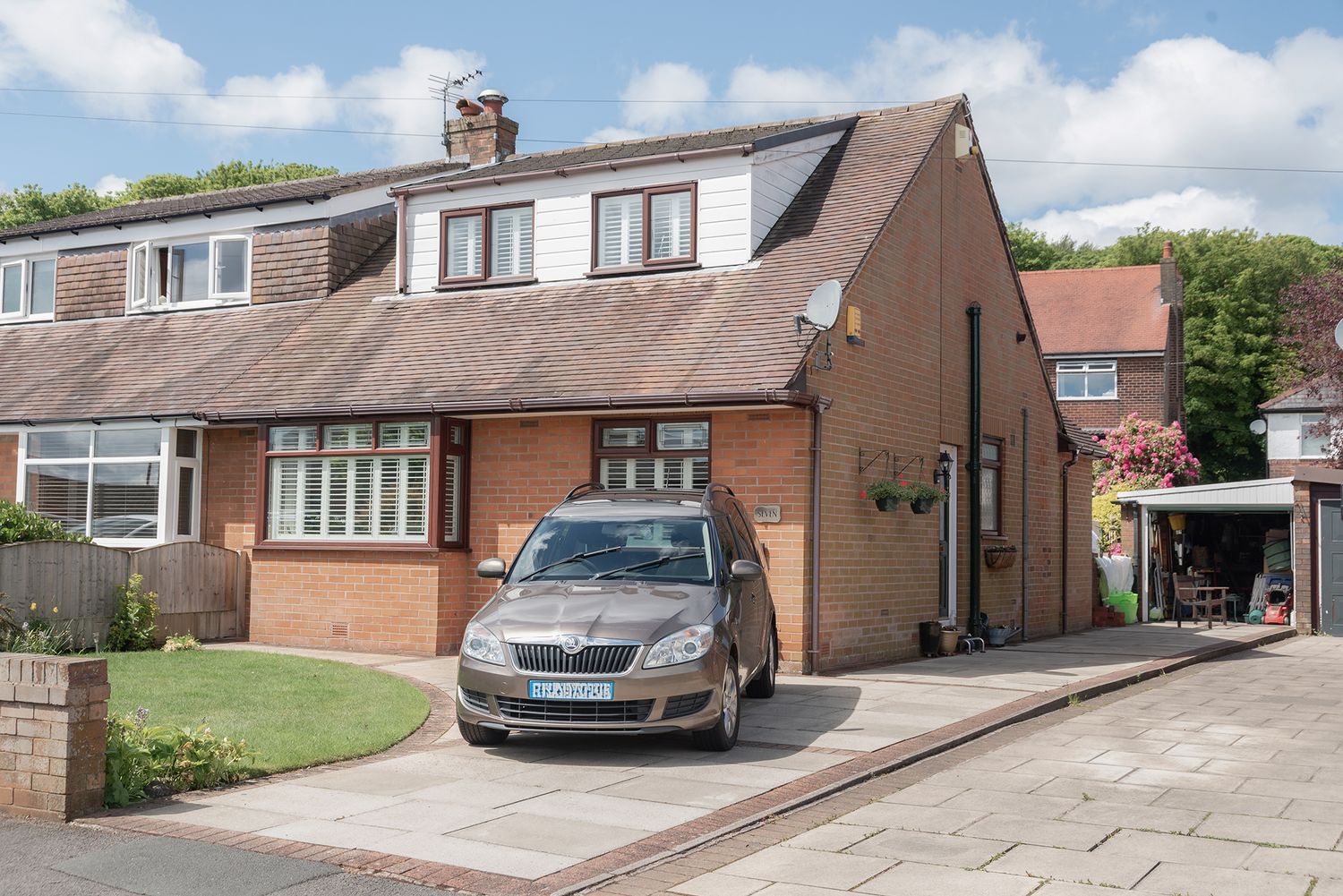 Semi-detached brick house with a grey car parked on a block-paved driveway, featuring a dormer roof and well-maintained front garden.
