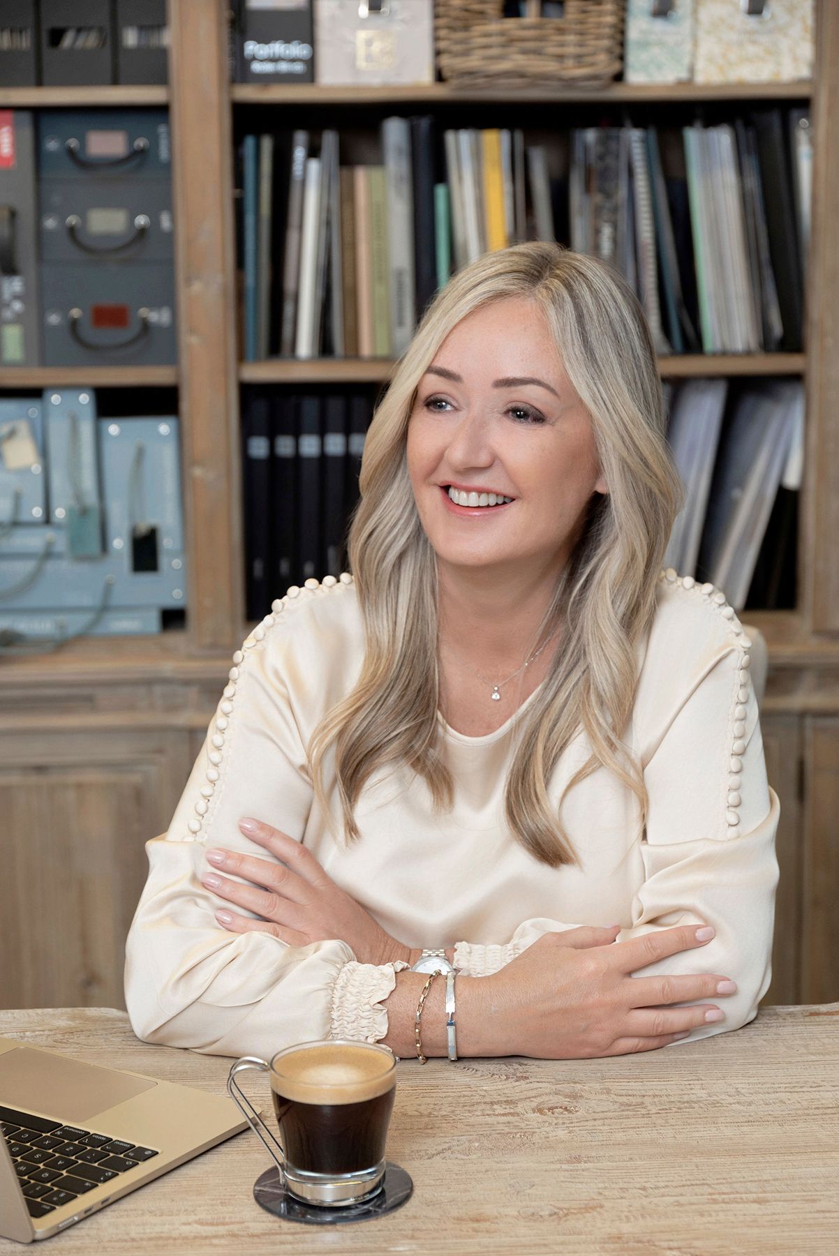 Smiling blonde woman in cream blouse sits at wooden desk with coffee and laptop, bookshelf behind her.