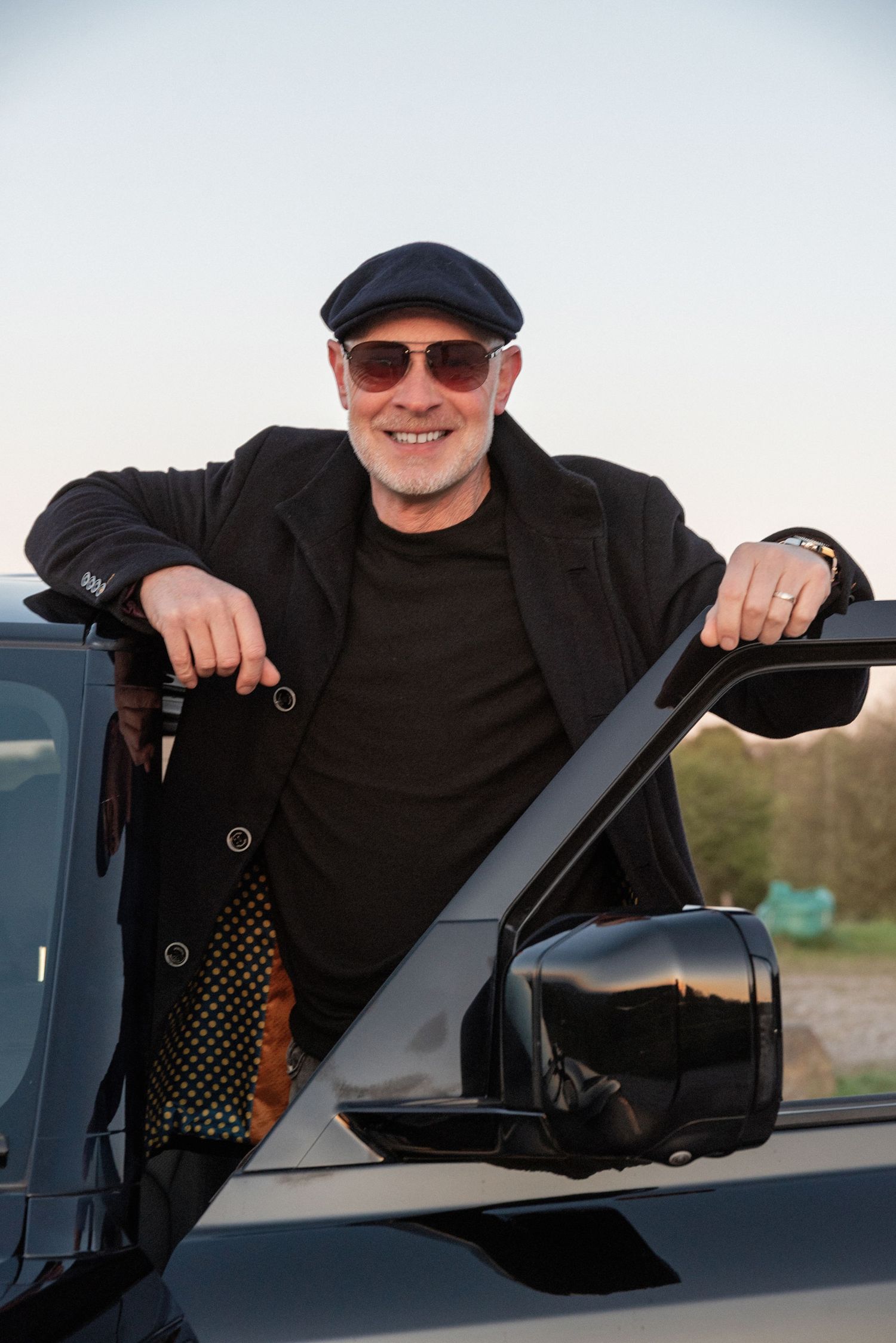 Smiling middle-aged man in black outfit, flat cap, and sunglasses leaning on open car door outdoors.