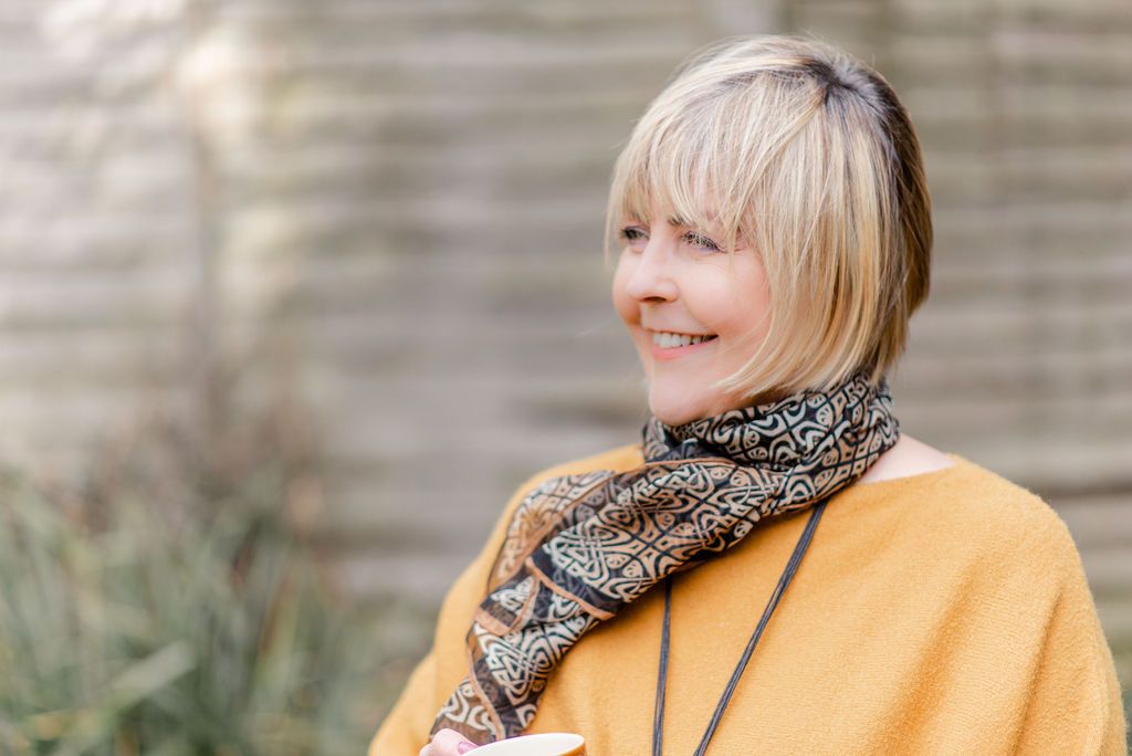 Smiling blonde woman with short bangs wearing a mustard yellow sweater and patterned scarf outdoors.