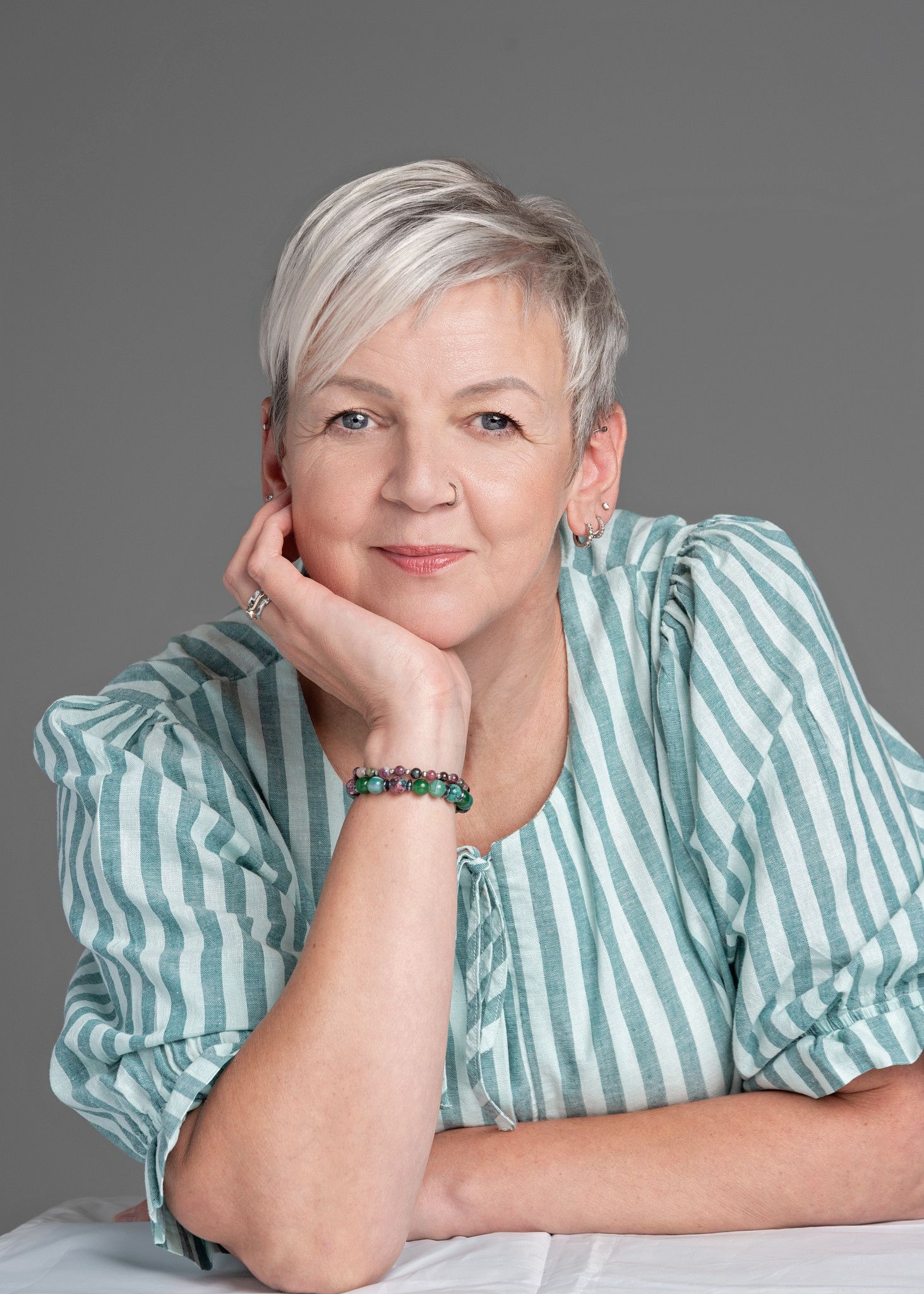 Smiling middle-aged woman with short gray hair wearing a green striped blouse, resting chin on hand against gray background.