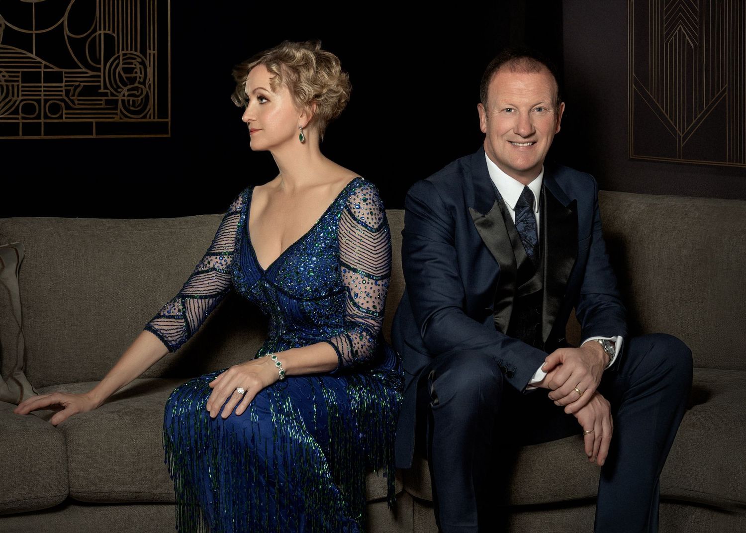 Couple in formal evening wear pose together on grey couch with art deco background.