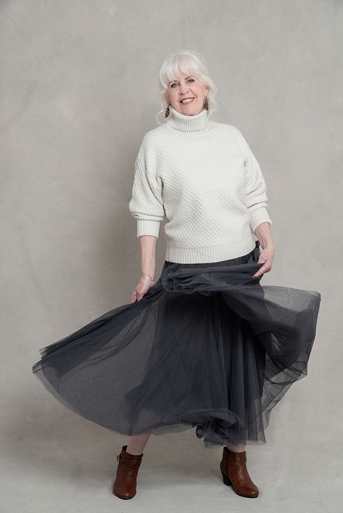 Woman in white turtleneck sweater twirling in flowing black tulle skirt and brown boots.