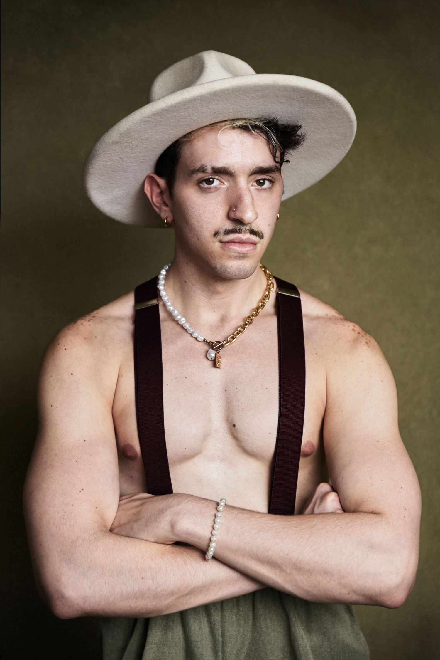 Dramatic portrait of a shirtless model in suspenders and white hat with jewelry against moody backdrop.