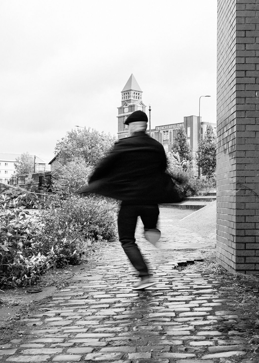 Blurred figure running on cobblestone path past brick wall with church tower in black and white urban scene.