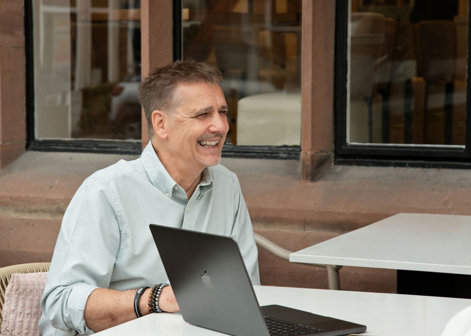 Smiling middle-aged man working on laptop outdoors at cafe table, wearing light blue shirt and bracelet.