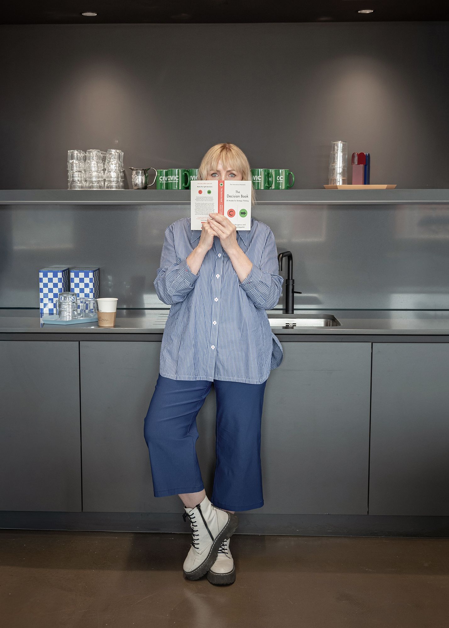Woman in blue striped shirt and wide-leg pants holds book in modern kitchen with gray cabinets.