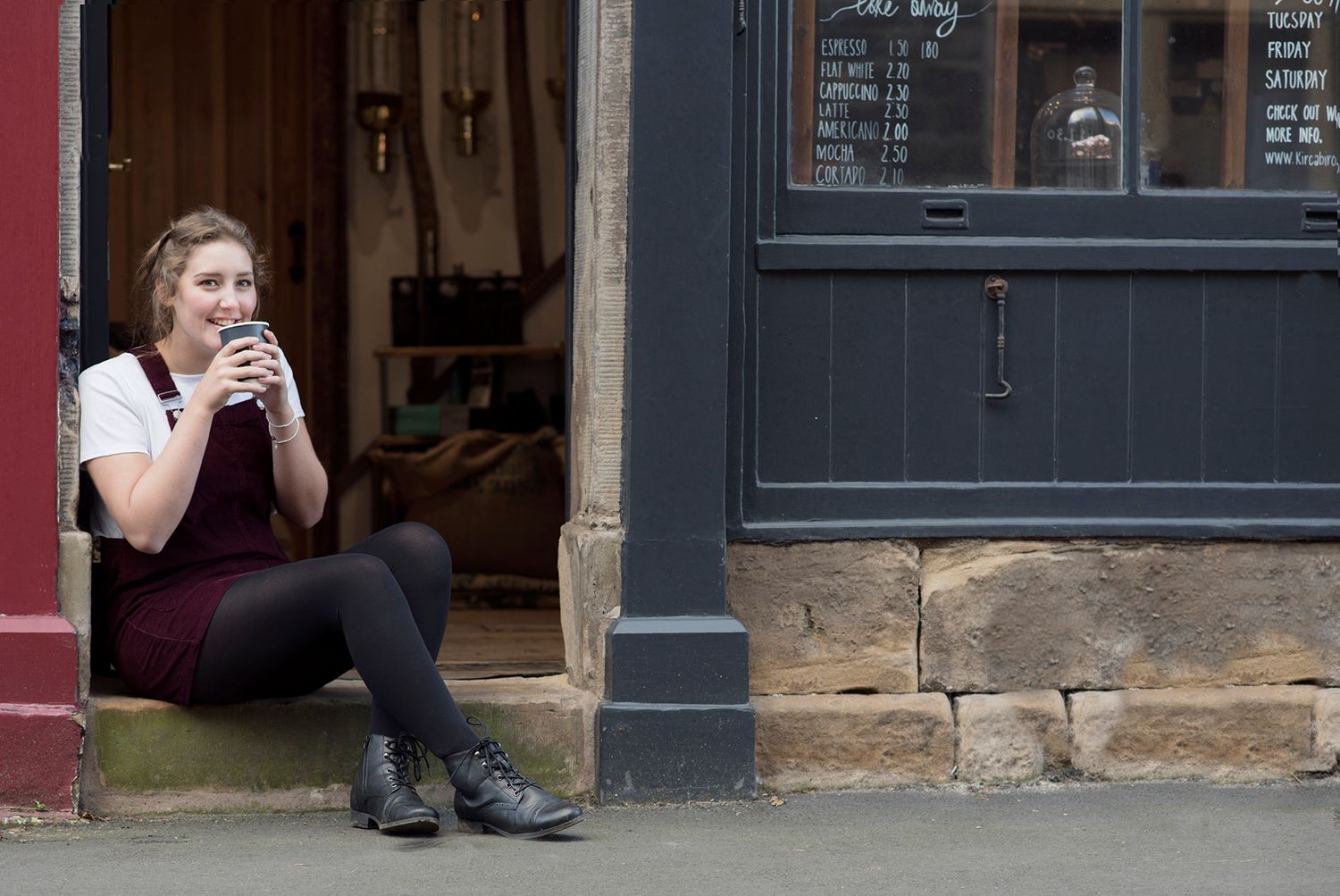 Young woman in apron sitting in shop doorway enjoying a hot drink outdoors.