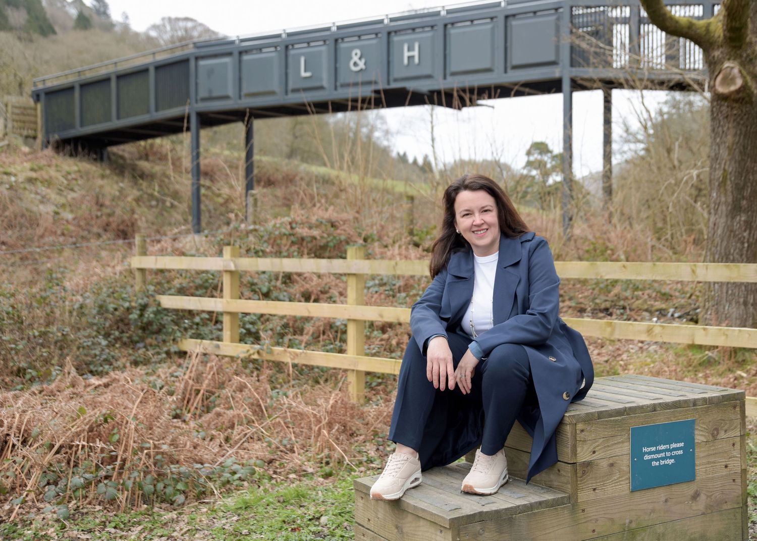 A person sits on a wooden bench beneath a large metal railway bridge in a rural hillside setting.