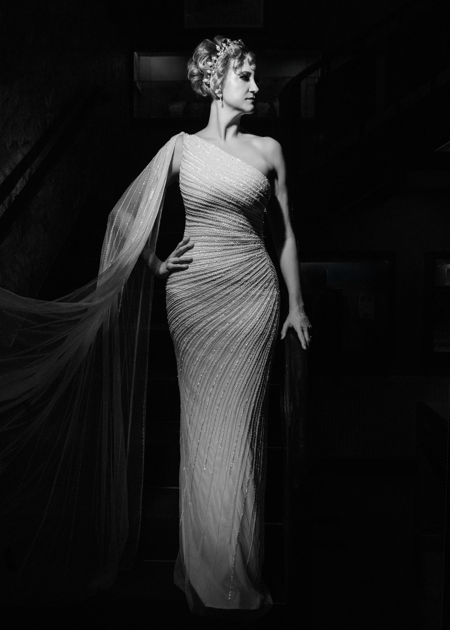 Black and white portrait of bride in fitted one-shoulder gown with flowing cape detail.