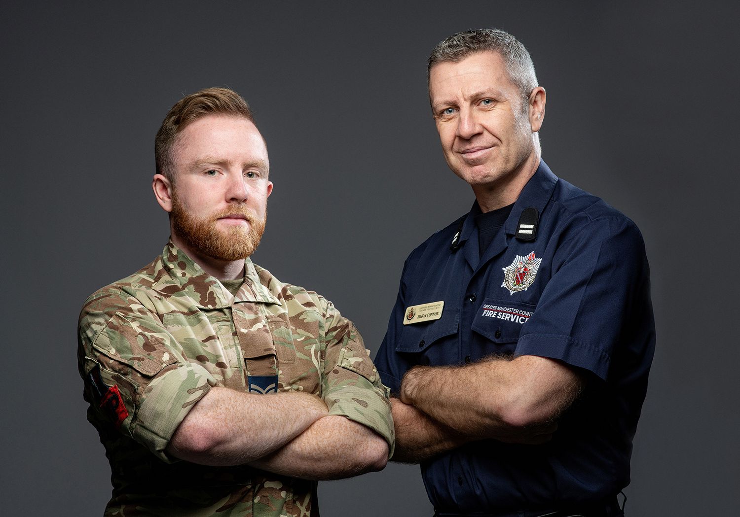 Two men in military uniforms, one in camouflage and one in navy blue, pose with crossed arms against a dark background.