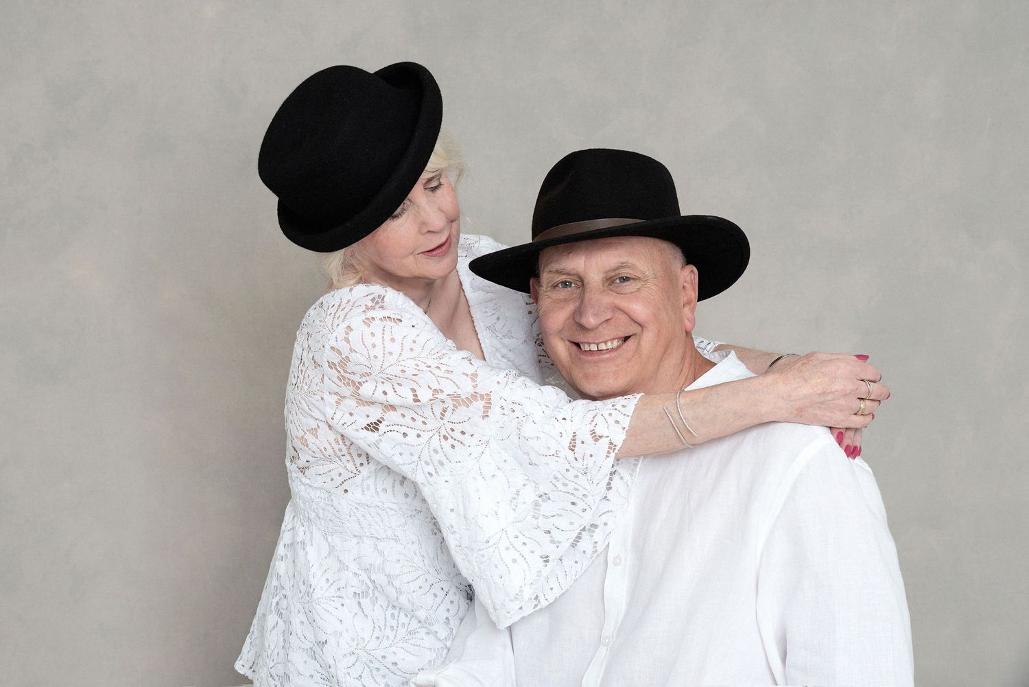 Couple in white attire and black hats embrace in a playful studio portrait.