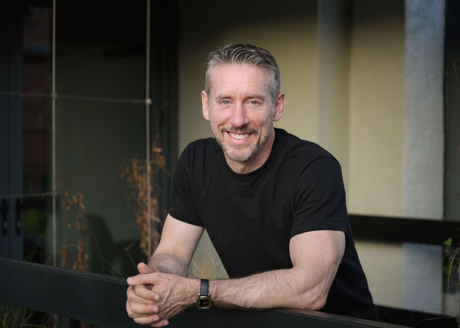 Smiling middle-aged man with gray hair wearing a black t-shirt, leaning on a railing outdoors.