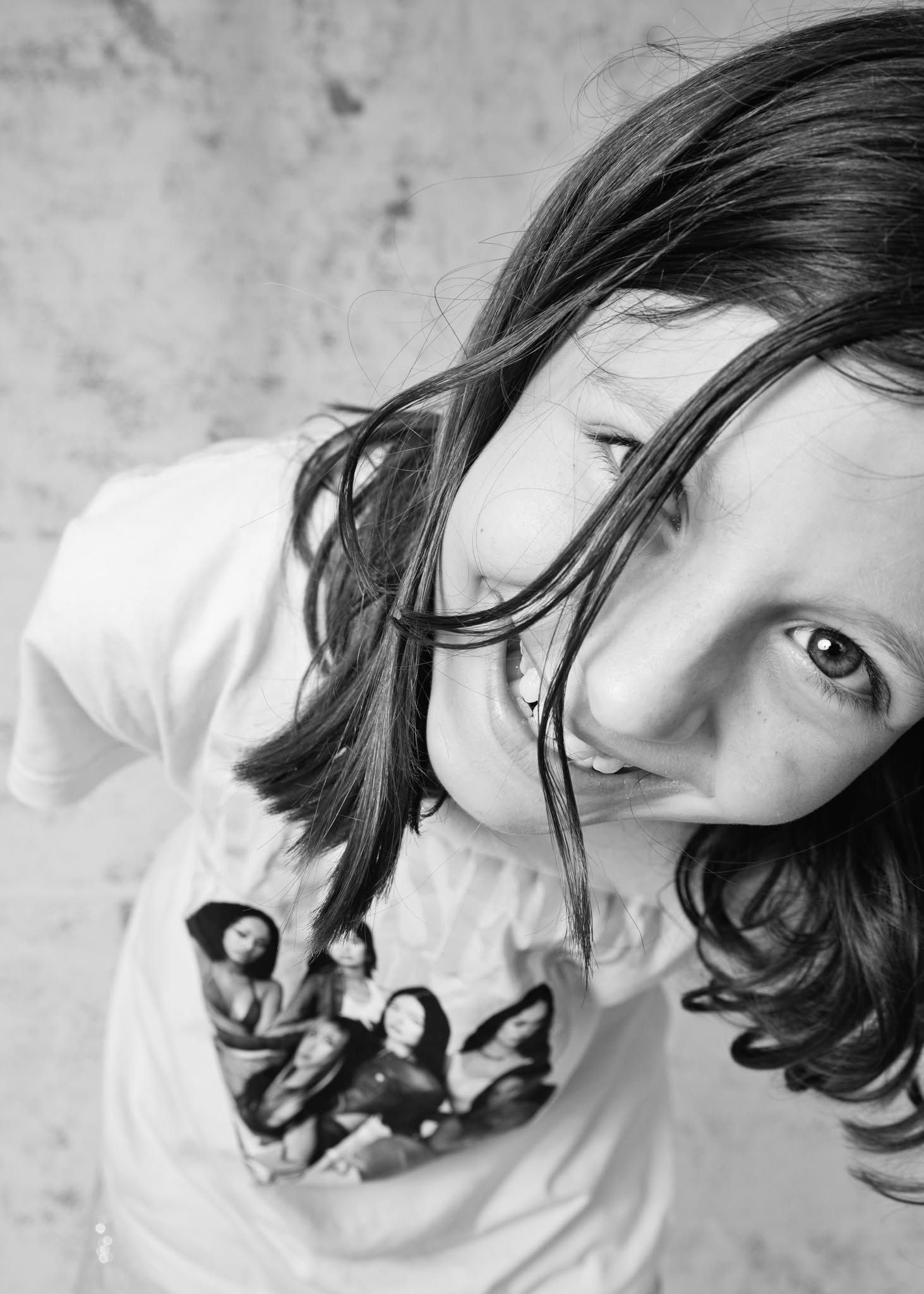 Black and white photo of a young girl with long hair looking up at camera, wearing a white graphic t-shirt.