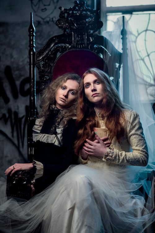 Two women in vintage Victorian costumes sit together on an ornate throne in an abandoned graffiti room.