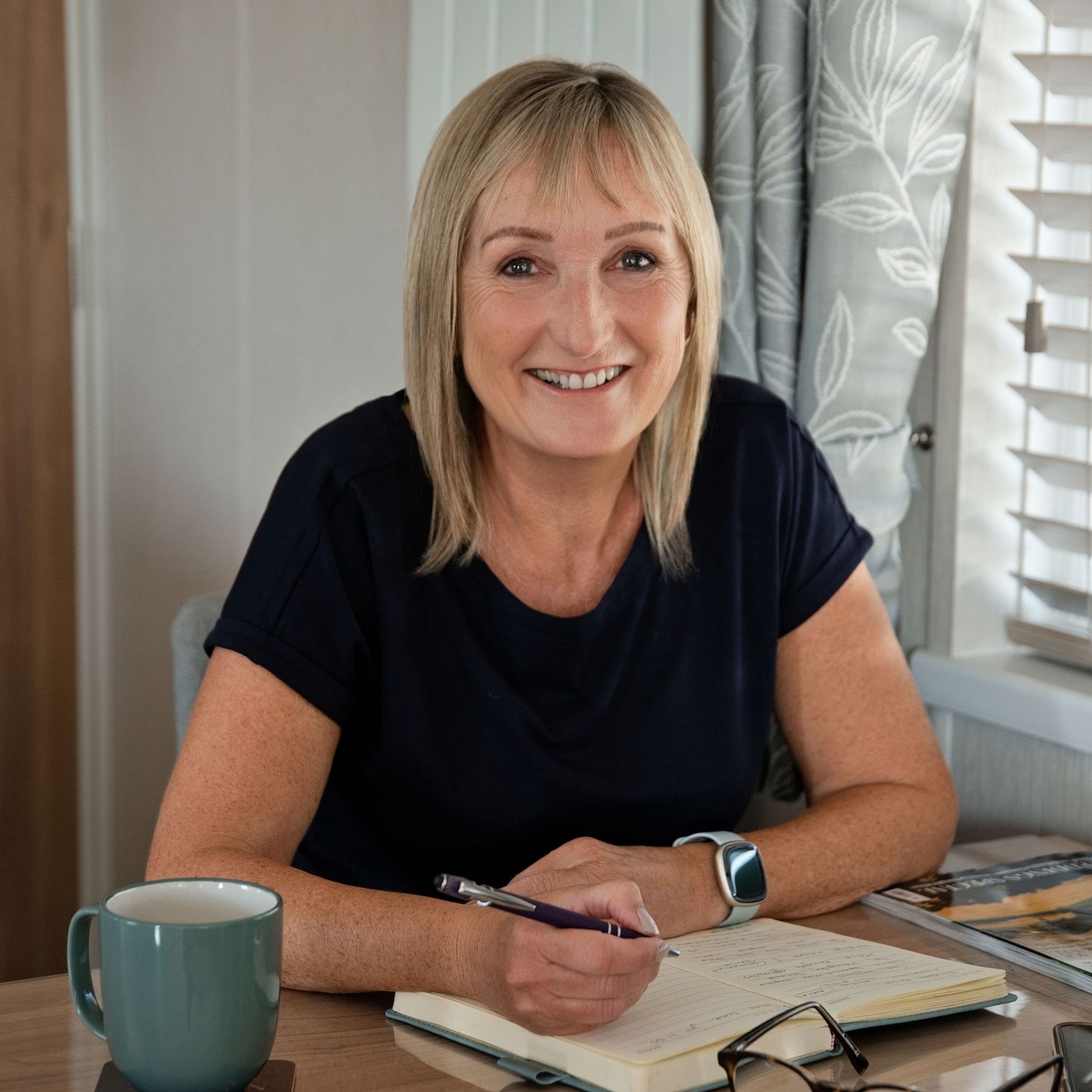 Smiling blonde woman writing in a notebook at a wooden desk with a teal mug and glasses nearby.