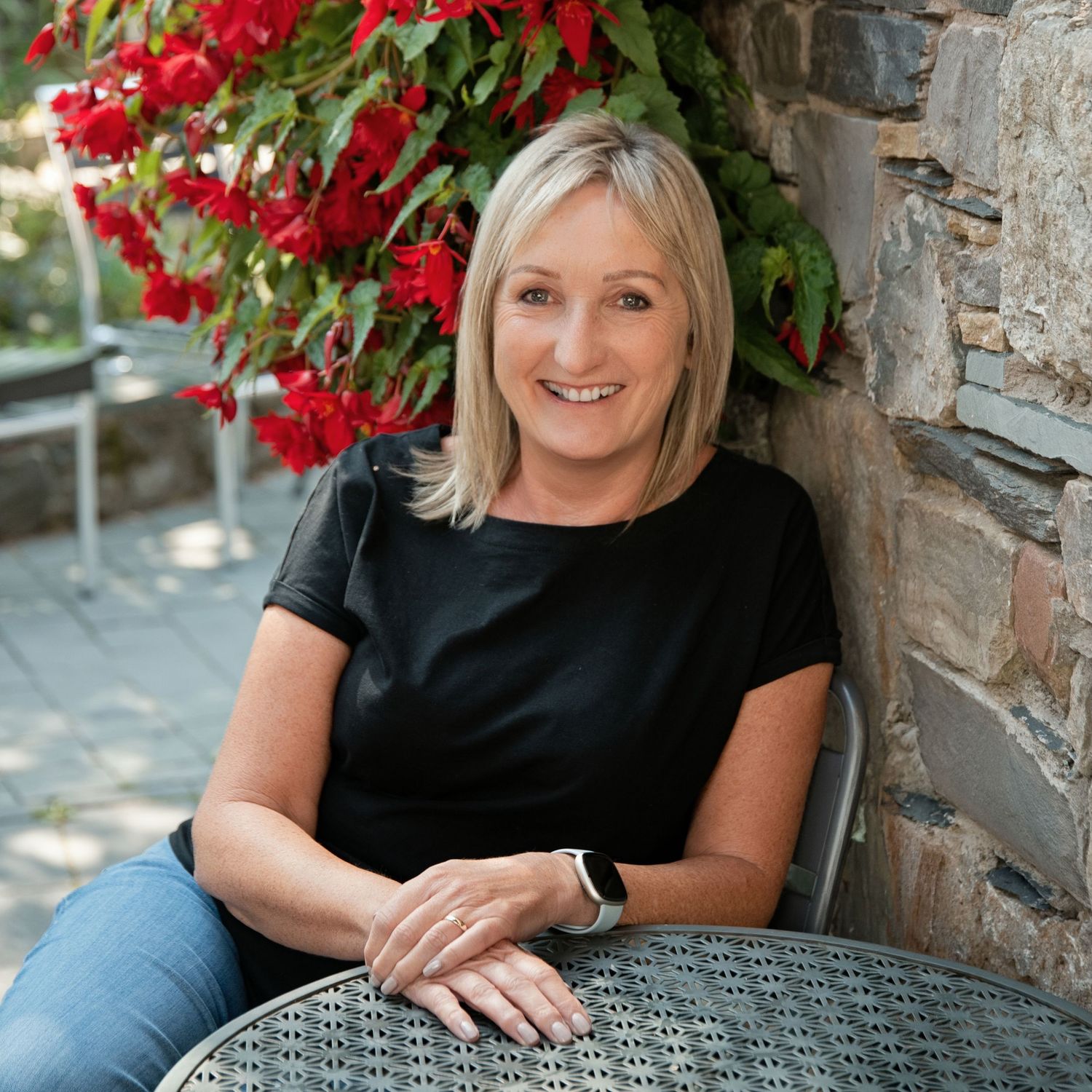 Smiling blonde woman in black top and jeans seated outdoors by stone wall with red flowers.