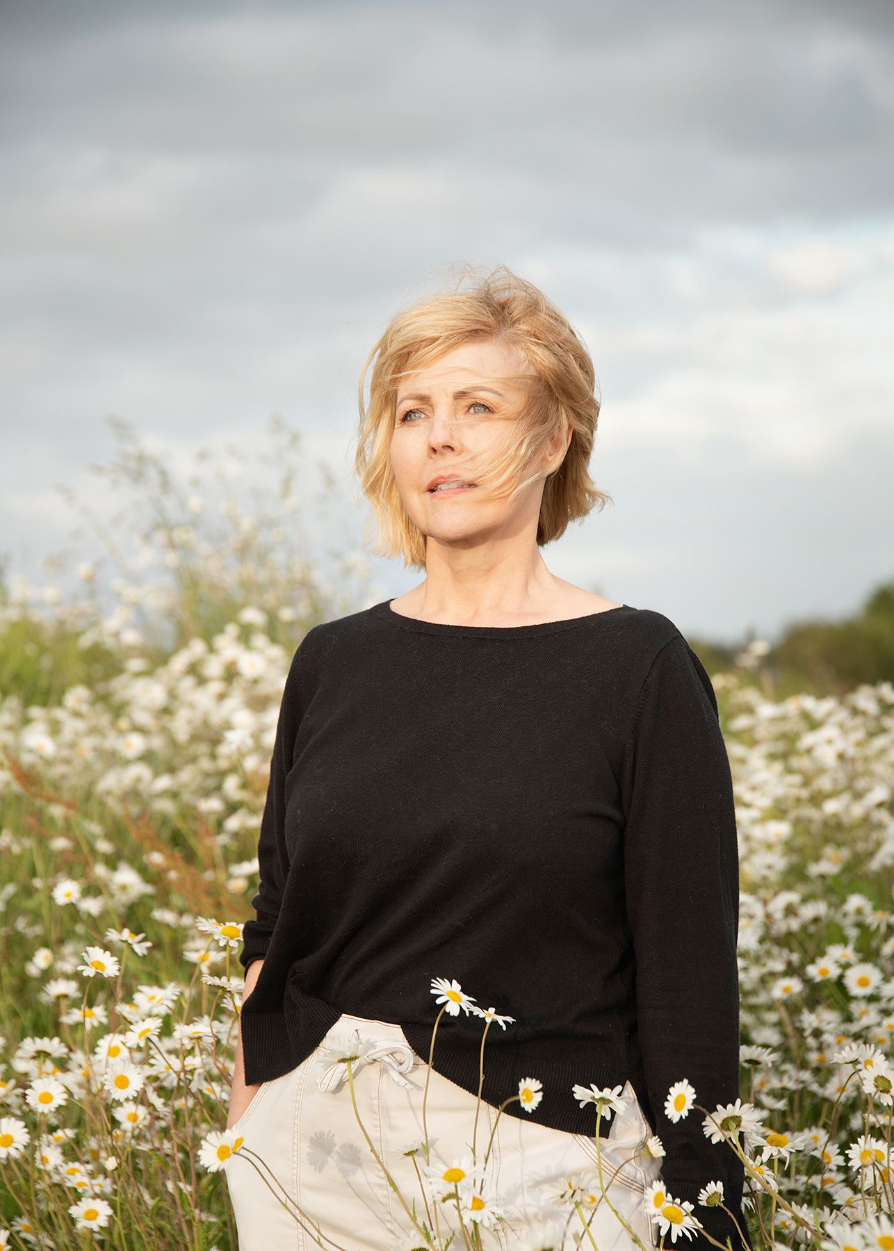 A middle-aged blonde woman in a black top stands among white daisies in a field under a cloudy sky.